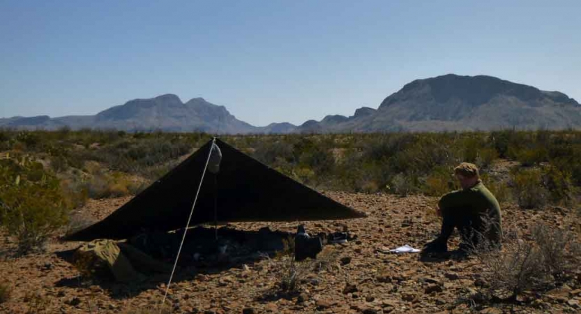 a person sits beside a tarp shelter in the desert with mountains in the background